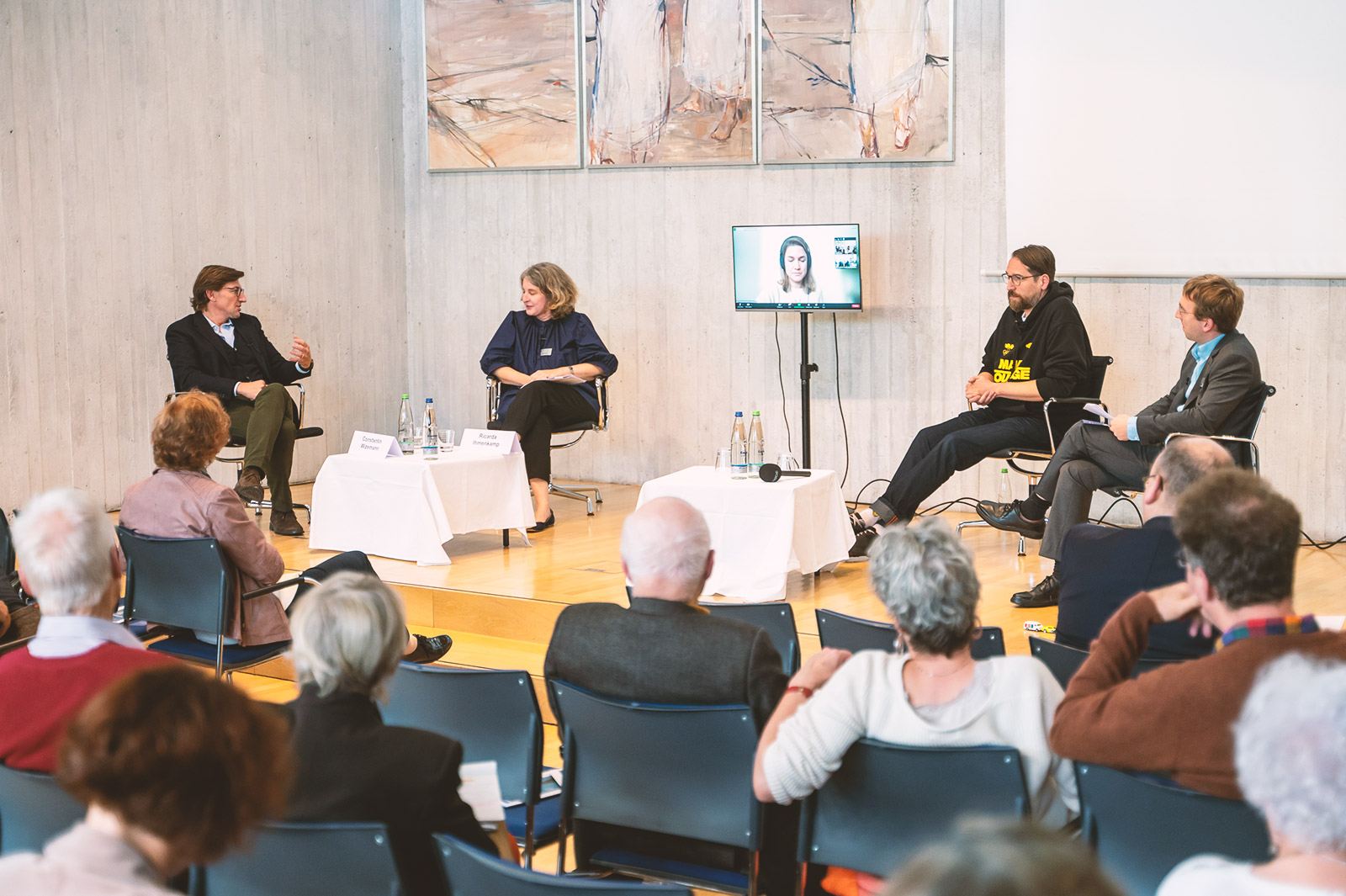 Podiumsdiskussion im Festsaal der Evangelischen Akademie Bad Boll. Vier Personen auf dem Podium. Eine Person auf Bildschirm zugeschaltet.